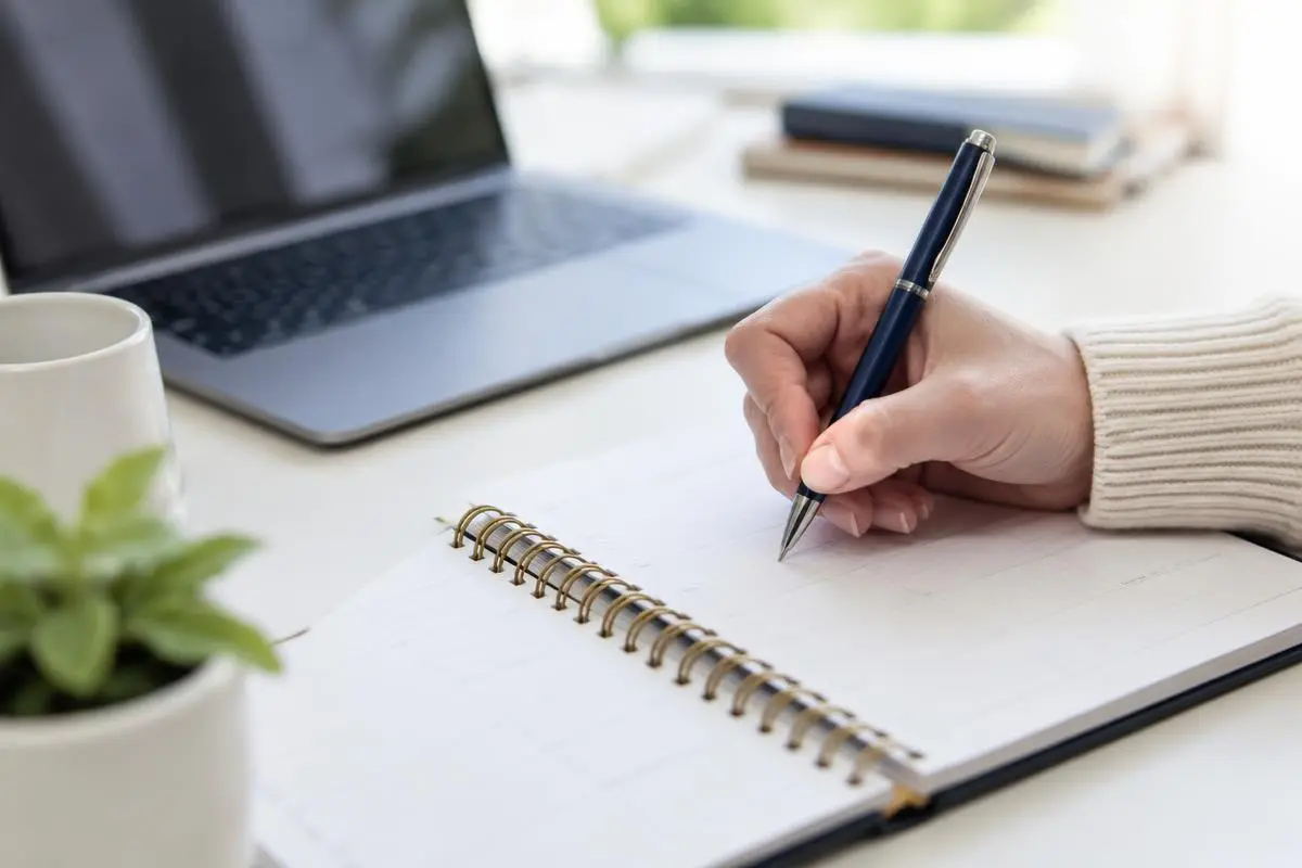 Hand writing goals in a planner, laptop in background