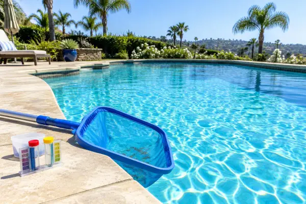 Pool skimmer net and water test kit beside a clear backyard pool